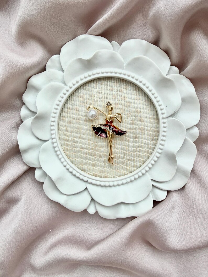 May include: A gold-toned ballerina brooch featuring a dark red skirt with clear crystals and a single white pearl accent, mounted inside a circular white frame with a scalloped edge and a textured beige fabric backing.