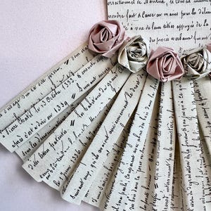 May include: A fan-shaped arrangement of paper strips with handwritten text in French. The strips are decorated with pink and white fabric roses.
