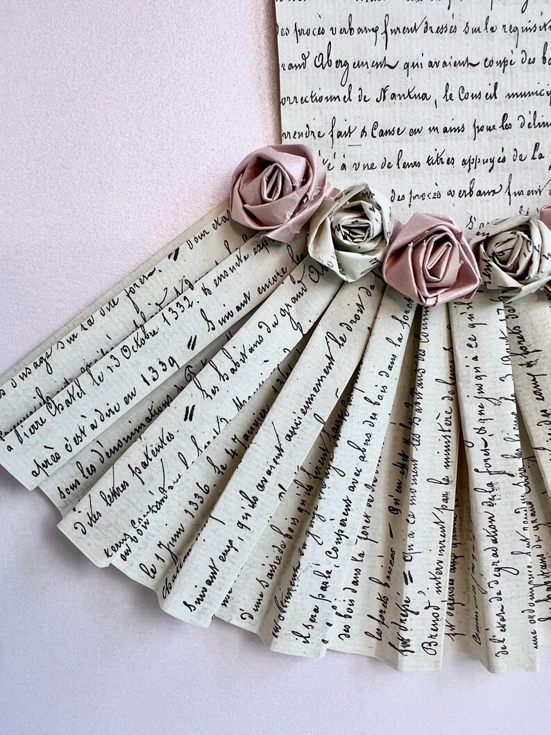 May include: A fan-shaped arrangement of paper strips with handwritten text in French. The strips are decorated with pink and white fabric roses.
