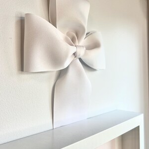 May include: A large white bow made of satin ribbon is attached to a white wall. The bow is tied in a simple knot and the ends of the ribbon are left long.