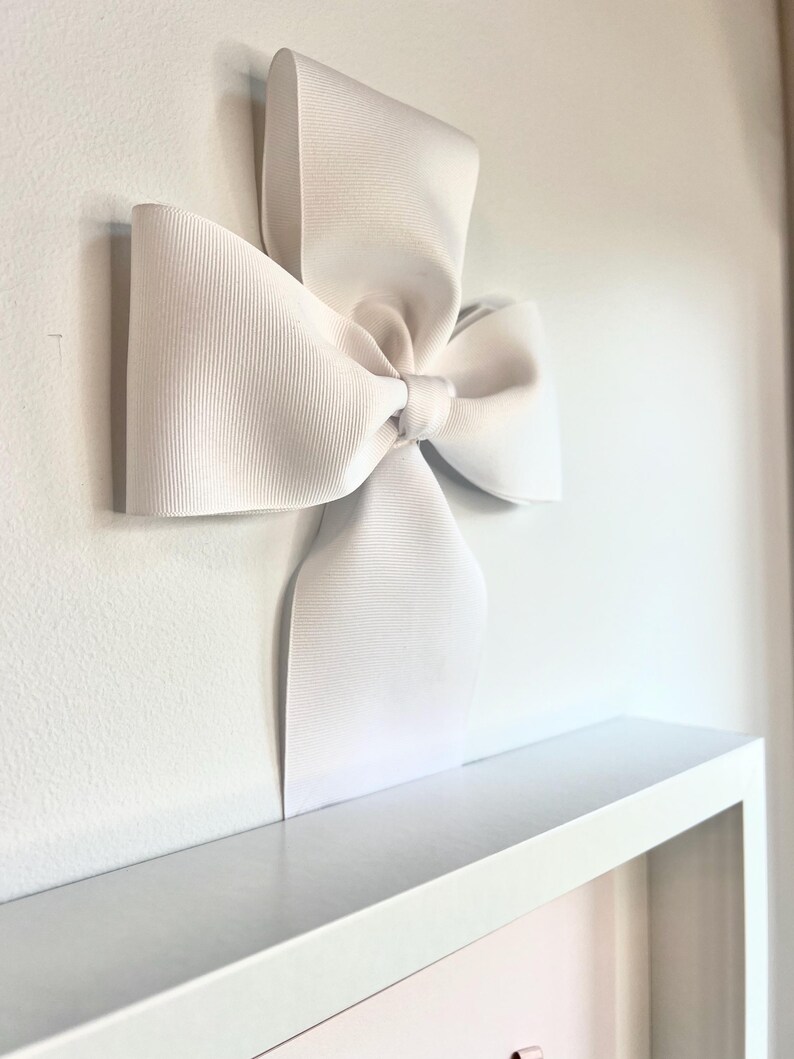 May include: A large white bow made of satin ribbon is attached to a white wall. The bow is tied in a simple knot and the ends of the ribbon are left long.