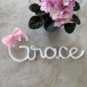 May include: White crocheted word "Grace" with a pink bow on a light gray surface. A potted pink African violet plant is in the background.