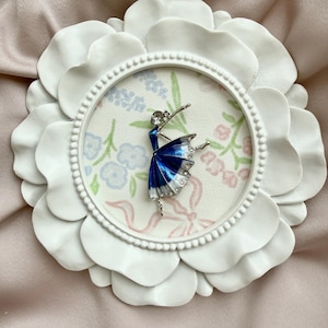 May include: A silver-toned ballerina brooch featuring a deep blue and white enamel skirt with clear rhinestone accents, displayed inside a circular white frame with a floral patterned background.