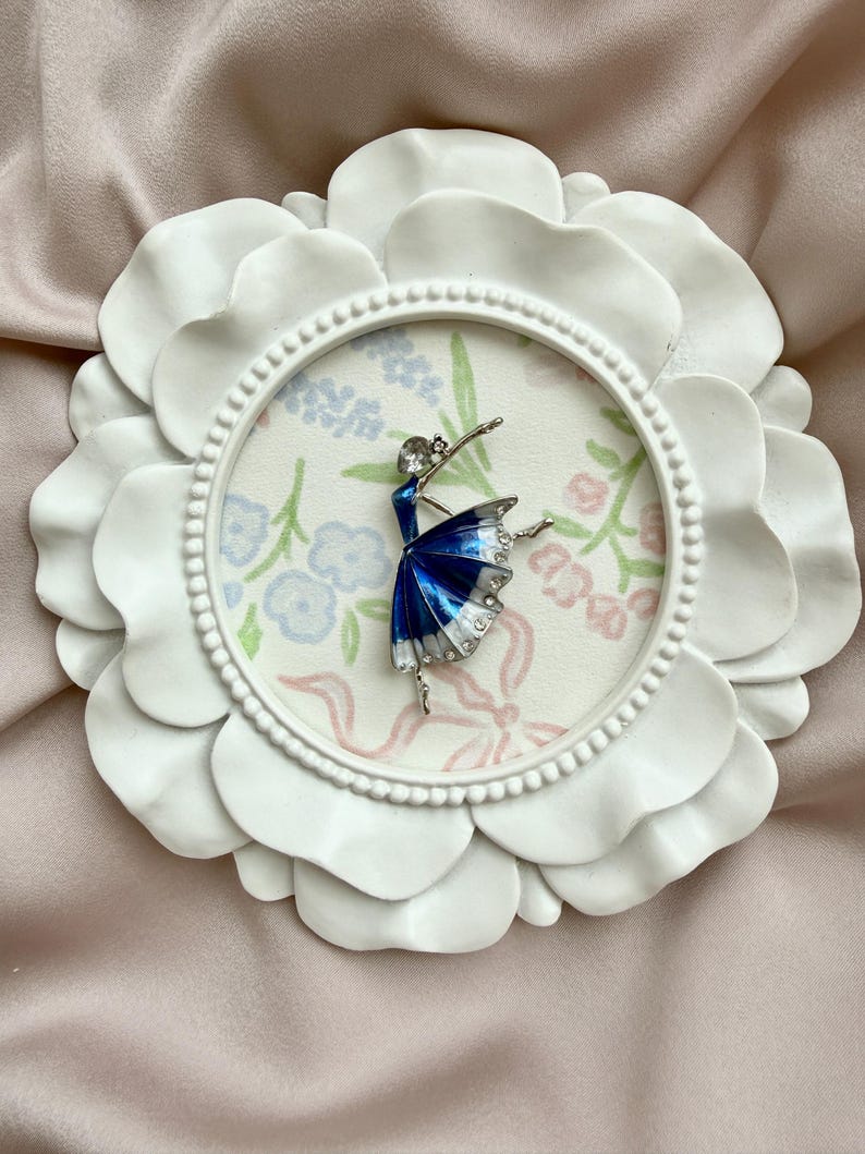 May include: A silver-toned ballerina brooch featuring a deep blue and white enamel skirt with clear rhinestone accents, displayed inside a circular white frame with a floral patterned background.