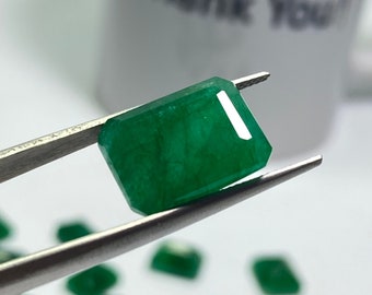 May include: A vibrant green emerald gemstone held in tweezers. The emerald is rectangular in shape and has a smooth, polished surface.