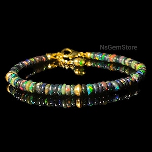Puede incluir: Una pulsera de cuentas con pequeñas piedras preciosas de ópalo negro redondas con destellos de color. La pulsera tiene detalles de metal dorado y un cierre. Las piedras preciosas están ensartadas, creando una joya delicada.
