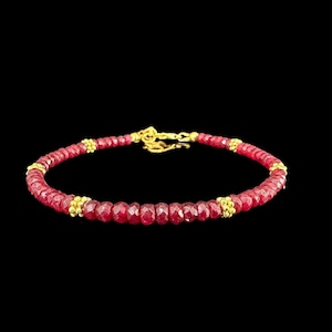 Könnte beinhalten: Ein Armband mit einer durchgehenden Reihe kleiner, facettierter, tiefroter Perlen. Das Design wird durch vier goldfarbene Metallzwischenstücke mit einer texturierten, körnigen Oberfläche unterbrochen. Ein goldfarbener Karabinerverschluss.