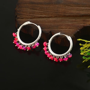 May include: A pair of silver hoop earrings featuring small, dangling, vibrant pink beads attached along the lower curve of each hoop. The earrings are circular in shape with a hinged closure mechanism, showcasing a polished metallic finish.