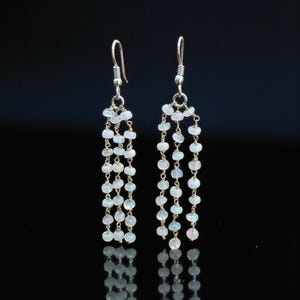 May include: A pair of dangling earrings with a cascade of small, round, white beads. The earrings have silver-colored hooks and chains. The beads have a subtle iridescent quality, reflecting light. The background is black.