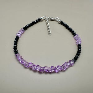 May include: A delicate bracelet featuring alternating sections of black and light purple beads. The bracelet has a silver-colored clasp and extender chain. The light purple beads are irregularly shaped, while the black beads are faceted.