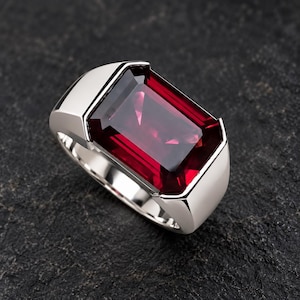 May include: A silver-toned ring featuring a large, rectangular, faceted deep red gemstone set in a smooth, polished metal band. The stone displays internal reflections and a vibrant crimson hue, securely held within a wide, solid metal setting.