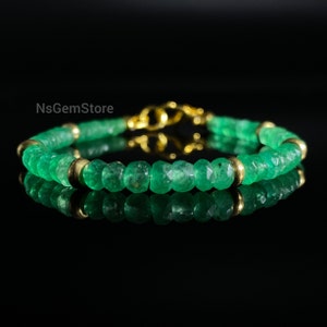 Puede incluir: Un brazalete de piedras preciosas de esmeralda verde con cuentas doradas y un cierre dorado. El brazalete está sobre un fondo negro.