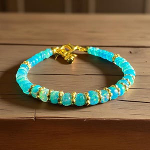 May include: A beaded bracelet featuring light blue and gold accents. The bracelet is composed of small, round, light blue beads interspersed with gold-colored spacers. The clasp and connecting elements are also gold. The bracelet is displayed on a wooden surface.