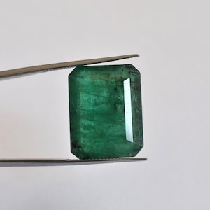 May include: A rectangular emerald gemstone with a deep green hue and visible internal inclusions, held by a pair of metal tweezers. The stone features a faceted cut with sharp corners and a polished surface that reflects light across its structure.