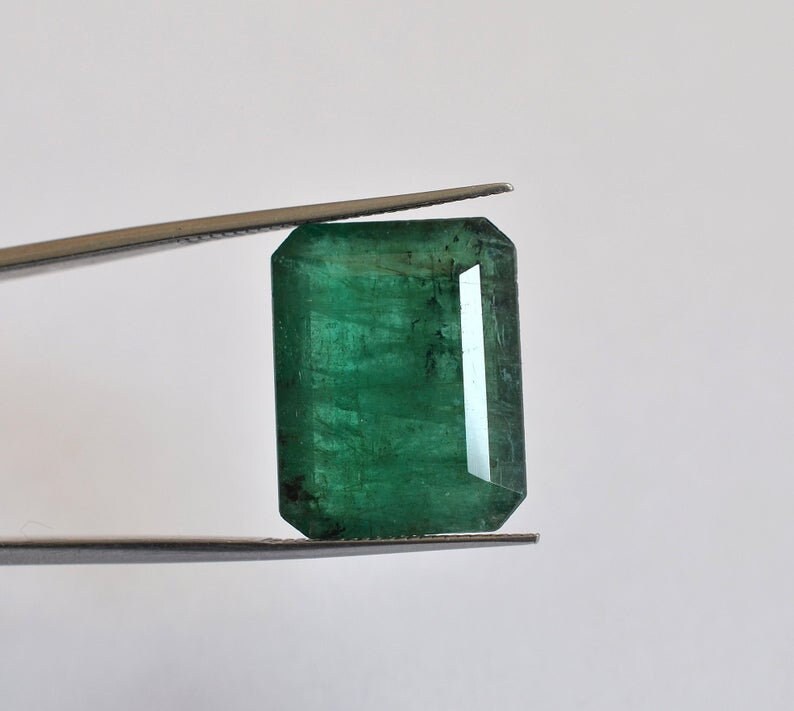May include: A rectangular emerald gemstone with a deep green hue and visible internal inclusions, held by a pair of metal tweezers. The stone features a faceted cut with sharp corners and a polished surface that reflects light across its structure.
