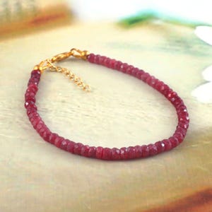 May include: A delicate bracelet composed of small, faceted, deep red gemstone beads strung together. The piece features a gold-toned metal lobster claw clasp and a short extender chain, providing an adjustable fit for the wearer's wrist.
