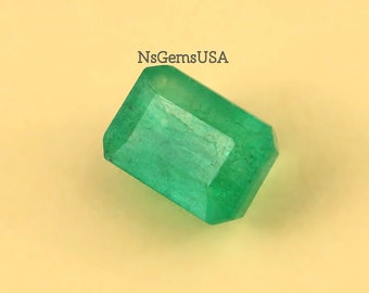 May include: A rectangular, faceted emerald gemstone with a vibrant green hue. The gem is set against a light yellow background. The text "NsGemsUSA" is visible in the upper right corner.