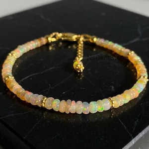 May include: A delicate bracelet featuring small, round, iridescent opal beads with a play of colour, interspersed with tiny gold-toned metal spacer beads. The bracelet includes a gold-toned chain extension with a small charm and the text NsGemStore.