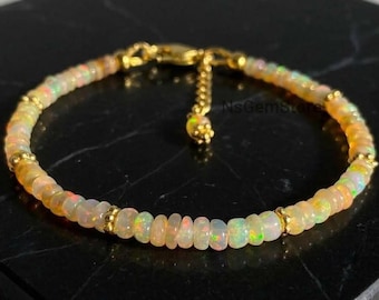 May include: A delicate bracelet featuring small, round, iridescent opal beads with a play of color, interspersed with tiny gold-toned metal spacer beads. The bracelet includes a gold-toned chain extension with a small charm and the text NsGemStore.