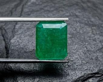 May include: A vibrant green emerald gemstone with a rectangular step-cut facet pattern. The gemstone is held securely by a pair of thin, silver-toned metal tweezers, showcasing its translucent quality and deep, saturated forest green color.