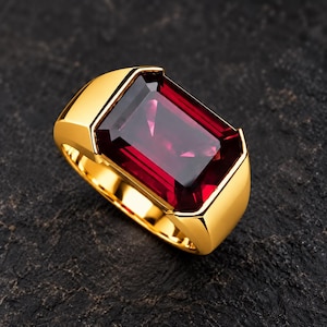 May include: A gold ring featuring a large, rectangular, deep red gemstone set in a polished yellow gold band. The stone has a faceted cut that reflects light, and the metal band has a smooth, solid finish with a thick, sturdy construction.