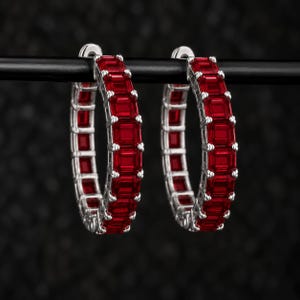 May include: A pair of hoop earrings crafted from silver-toned metal, each featuring a continuous row of vibrant red, emerald-cut gemstones set in a prong setting. The circular design showcases a uniform arrangement of stones around the entire hoop.