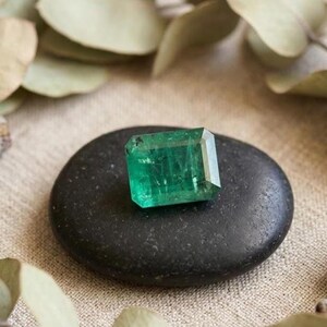 May include: A rectangular emerald-cut gemstone with a vibrant green hue and faceted surfaces, resting on a smooth, dark charcoal-colored oval stone. The gemstone displays internal clarity characteristics and reflects light across its top facets.