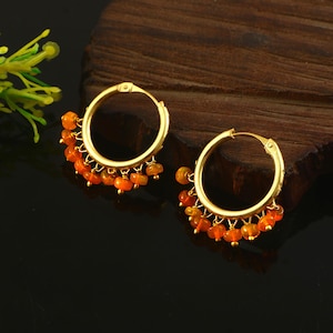 May include: A pair of gold hoop earrings featuring a series of small, round, vibrant orange beads dangling from the bottom half of each hoop. The earrings have a polished metallic finish and a secure clasp mechanism at the top of each circular frame.