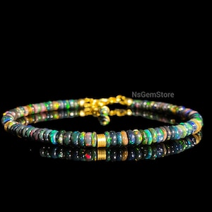 Peut inclure: Un bracelet de perles composé de petites perles rondes d'opale noire avec des éclats de couleurs. Le bracelet a des accents dorés et un fermoir. Le bijou est présenté sur une surface réfléchissante sur fond noir.