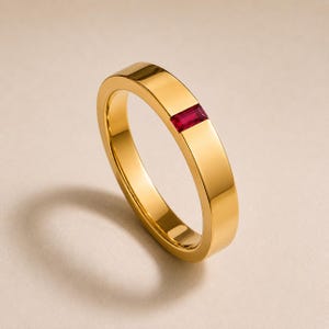 Op de afbeelding: Een gepolijste gouden ring met een enkele rechthoekige, in baguette geslepen rode edelsteen die verzonken in het midden van het metalen oppervlak is geplaatst. De ring heeft een gladde, reflecterende afwerking en een constante breedte.