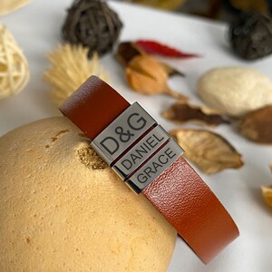Puede incluir: Un brazalete de cuero marr&oacute;n con tres abalorios de metal plateado. Los abalorios est&aacute;n grabados con los nombres "D&G", "Daniel" y "Grace".