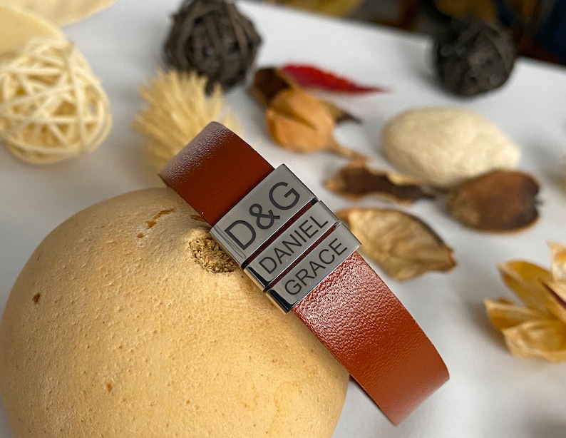 Puede incluir: Un brazalete de cuero marr&oacute;n con tres abalorios de metal plateado. Los abalorios est&aacute;n grabados con los nombres "D&G", "Daniel" y "Grace".
