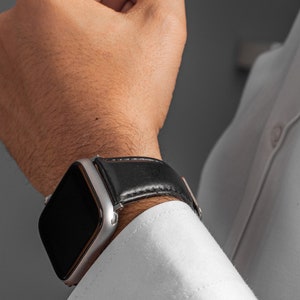 May include: A silver smartwatch with a black leather strap on a person's wrist.