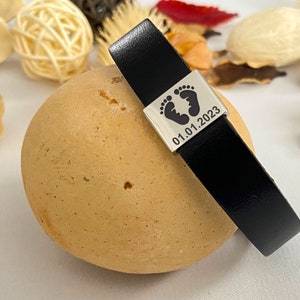 Puede incluir: Pulsera de cuero negro con un dije de plata con un dise&ntilde;o de huella y la fecha "01.01.2023".