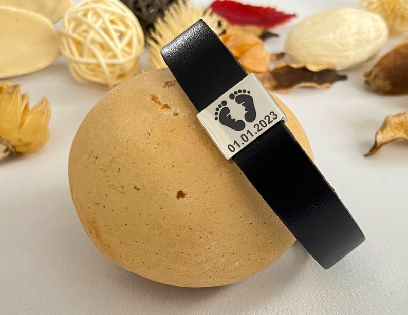 Puede incluir: Pulsera de cuero negro con un dije de plata con un dise&ntilde;o de huella y la fecha "01.01.2023".