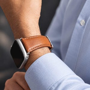 Peut inclure: Une montre connectée argentée dotée d'un bracelet en cuir marron clair avec des coutures apparentes. Le boîtier est rectangulaire aux coins arrondis avec un écran noir, fixé au poignet par une boucle en métal sur le bracelet en cuir.