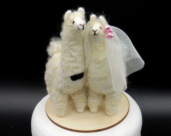 May include: White felt llama wedding cake topper with a black bow tie and a white veil with pink flowers. The llama couple is standing on a wooden base.