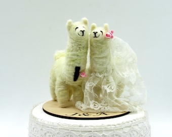 May include: A whimsical wedding cake topper featuring two felted llamas. One llama wears a black tie and a pink rose, the other a lace wedding dress and veil, with pink roses. The cake base is decorated with lace and a wooden disc with the initials "J & A".