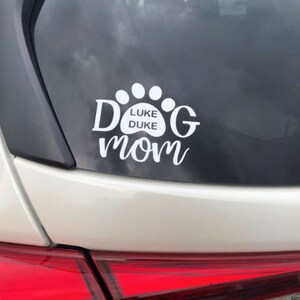May include: White vinyl car sticker with a paw print design. The paw print has the text "LUKE DUKE" inside. The text "DOG MOM" is below the paw print.