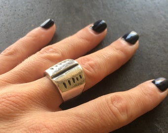 May include: A silver ring with a textured, rectangular design. The ring has a vertical line down the center and a series of horizontal lines on the side.