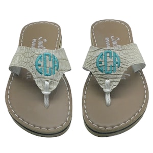 May include: A pair of beige flip-flops with a textured, crocodile-like pattern on the straps. Each strap features a teal monogram and the brand name. The soles are tan with a black and navy stripe.