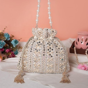 Puede incluir: Un bolso con cordón de color crema, adornado con cuentas, con un asa de perlas y borlas doradas. El bolso está adornado con pequeños espejos cuadrados y perlas, creando un efecto brillante. El asa está hecha de perlas blancas.