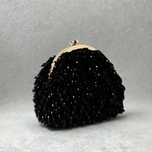 Könnte beinhalten: Eine schwarze, mit Perlen besetzte Clutch mit einem goldfarbenen Verschluss. Die Clutch ist mit kleinen, glänzenden schwarzen Perlen bedeckt, was ihr ein strukturiertes Aussehen verleiht. Der Verschluss ist aus abgerundetem, goldfarbenem Metall.