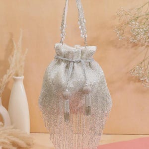 Puede incluir: Un bolso plateado con cuentas y cordón, con un asa blanca y detalles de cuentas. El bolso presenta una franja de cuentas colgantes y dos borlas decorativas. El bolso es de color plata claro y probablemente se utiliza para llevar objetos pequeños.