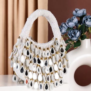 Può includere: Una borsa a mano bianca con perline con un design a forma di lacrima dorata e argentata. La borsa ha un'unica maniglia e una frangia di perline a forma di lacrima.