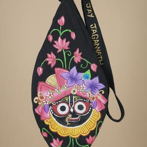May include: A black fabric shoulder bag featuring a hand-painted depiction of Lord Jagannath with large white eyes and a red face. The design includes pink lotus flowers and purple blossoms. The strap is embroidered with the text JAY JAGANATH.