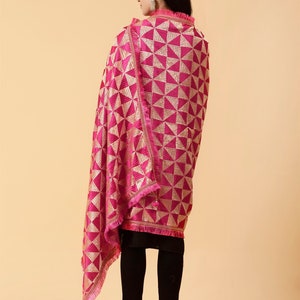 Może przedstawiać: R&oacute;żowy szal z wzorem w kolorze złotym i frędzlami. Szal jest zarzucony na ramiona osoby i noszony z czarnymi spodniami.