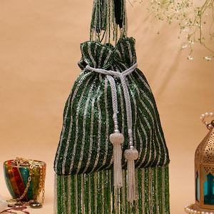 May include: A green and silver beaded drawstring bag with a long handle. The bag features vertical stripes and a beaded fringe along the bottom. Silver tassels hang from the drawstring closure. Decorative items are in the background.