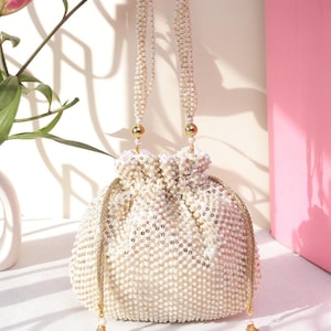 May include: Ivory beaded drawstring bag with pearl-like beads and gold accents. The bag has a round shape and a beaded handle. Tassels hang from the bottom. A pink lily and a white vase are in the background.