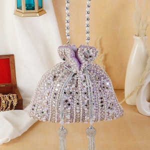 May include: A lavender beaded handbag with pearl and mirror embellishments. The bag has a drawstring closure and pearl beaded handles. Silver tassels hang from the bottom. A decorative lantern and vase are in the background.